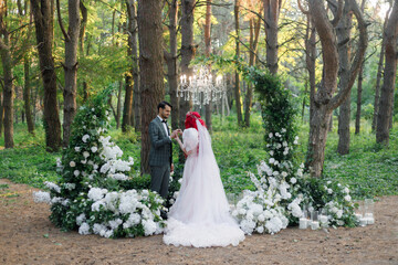 Beautiful couple, the groom in a green suit and the bride in an unusual pink wedding dress, posing at a luxurious ceremony with beautiful decor and an arch with a chandelier and candles in the forest 