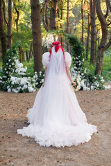 Beautiful couple, the groom in a green suit and the bride in an unusual pink wedding dress, posing at a luxurious ceremony with beautiful decor and an arch with a chandelier and candles in the forest 