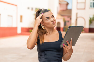 Young pretty woman holding a tablet at outdoors having doubts and with confuse face expression