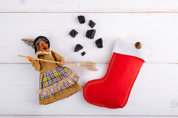 The witch Befana and red stocking with sweet coal and candy on white wooden background. Italian...