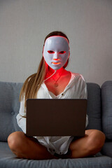 A beautiful girl with an LED mask on her head works at home on a laptop. Home skin care concept.