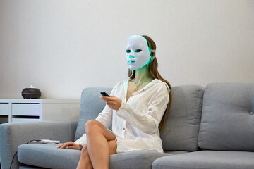 A beautiful girl with an LED mask on her head watches TV at home. Home skin care concept.