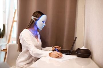 Naklejka premium A beautiful girl with an LED mask on her head works at a laptop. Home skin care concept.