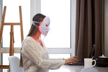 A beautiful girl with an LED mask on her head works at a laptop. Home skin care concept.