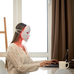 A beautiful girl with an LED mask on her head works at a laptop. Home skin care concept.