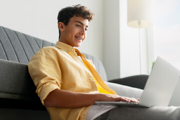 Smiling curly haired boy, teenager with dental braces on teeth using laptop computer studying,...