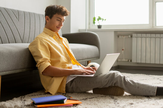 Attractive curly haired boy, teenager using laptop computer communication online sitting at home. Young student studying, learning language, exam preparation. Education concept 