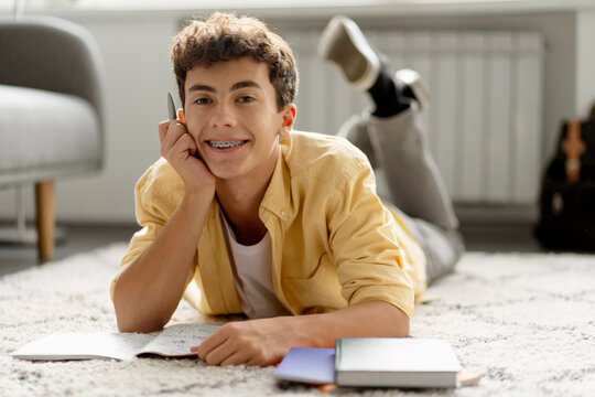 Smart smiling teenage boy with dental braces studying ta home, lying on floor looking at camera. Education concept