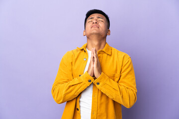 Young Ecuadorian man isolated on purple background keeps palm together. Person asks for something