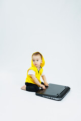 A cheerful 1-year-old boy in a yellow vest sits with a laptop on a white background. Modern...