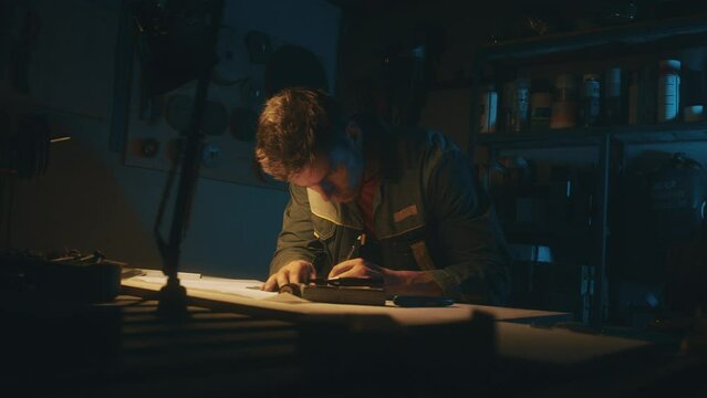 A metallurgical industry worker works on a prototype drawing on a steel table in a dark workshop with full equipment. Engineer working indoors on new product prototype in metal manufactory. 4K