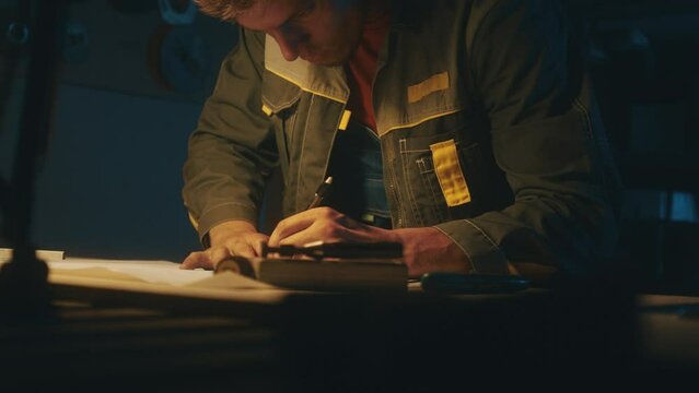 A metallurgical industry worker works on a prototype drawing on a steel table in a dark workshop with full equipment. Engineer working indoors on new product prototype in metal manufactory. 4K