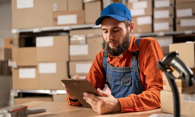 A warehouse worker conducting an inventory uses a digital tablet while sitting at a table in work clothes.