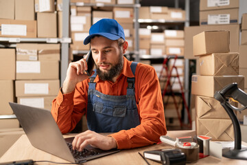 Male entrepreneur speaking on the phone while working on a laptop in warehouse. Online store owner...