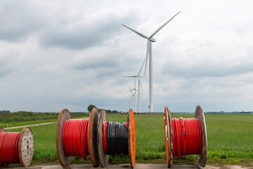 Number of large wheels or spools with black and red cable in front of a long row of modern wind...