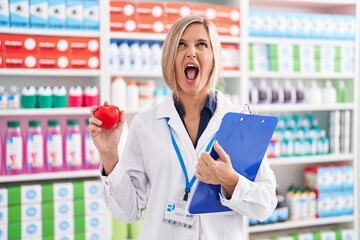 Young woman working at pharmacy drugstore holding heart angry and mad screaming frustrated and...