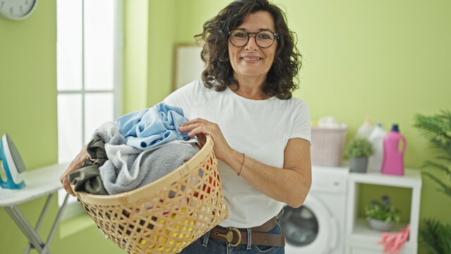 Middle age hispanic woman smiling confident holding basket with clothes at laundry room