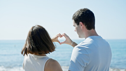 Beautiful couple doing heart gesture with hands standing backwards at seaside