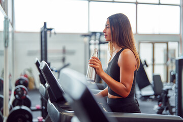 Fototapeta na wymiar Latin American woman walking on a treadmill and drinking water in the gym