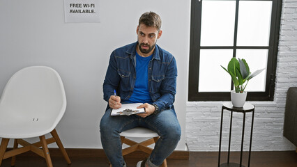 Handsome young hispanic man, a serious adult with a beard, relaxes in a chair, deeply immersed in writing a report on a clipboard in a quiet indoor waiting room setting. - obrazy, fototapety, plakaty