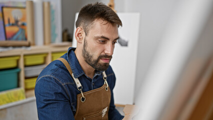 Relaxed young hispanic man, a portrait of handsome bearded artist, focused and concentrating on drawing with paintbrushes at art studio, perfecting canvas masterpiece