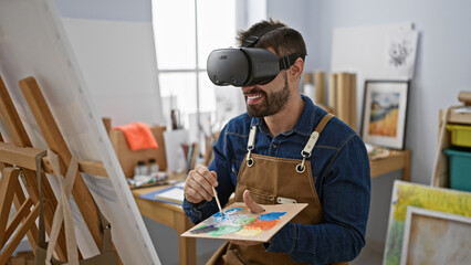 Charming young hispanic man artist smiling while drawing in high-tech virtual reality glasses at a...
