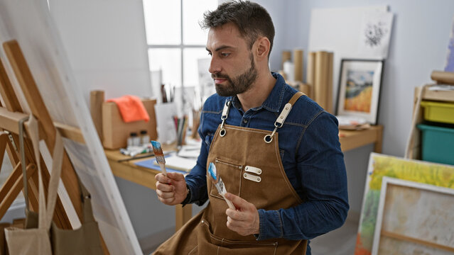 Young handsome hispanic man artist concentrates, draped in apron, beard highlighted, interiors of studio buzz with focus, drawing class at art academy, learning the brush strokes on canvas.