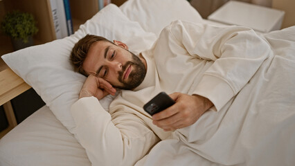 Handsome hispanic guy chilling at home, lying in bed, engrossed in texting on smartphone amidst a serene morning bedroom backdrop