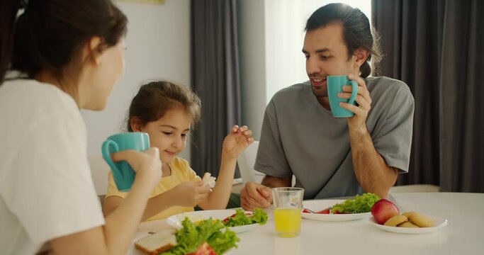 A brunette man in a gray T-shirt holds a turquoise cup in his hand and has breakfast with his wife and little daughter in a yellow dress with a white table in a modern kitchen