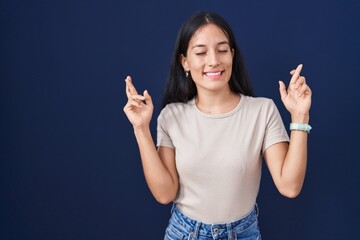 Young hispanic woman standing over blue background gesturing finger crossed smiling with hope and eyes closed. luck and superstitious concept.