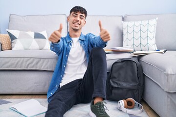 Young hispanic man sitting on the floor studying for university approving doing positive gesture...