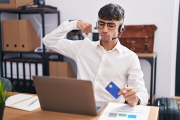 Young hispanic man working using computer laptop holding credit card strong person showing arm...