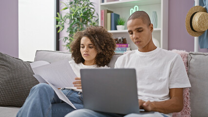 Beautiful couple in love, sitting together on their sofa at home, seriously concentrated on reading a bill on their laptop, embracing relaxed lifestyle indoors.