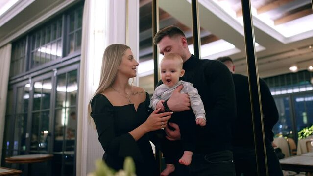 Smiling man holds his infant son waving him. Long-haired blonde woman takes a man by the arm and kisses baby.