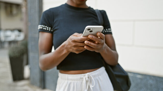 Attractive african american woman engrossed in digital conversation, using her phone outdoors, touching the screen to text in an urban city street setting.