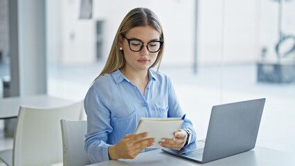 Young blonde woman business worker using laptop reading notebook at office