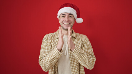 Young hispanic man wearing christmas hat clapping hands applause over isolated red background