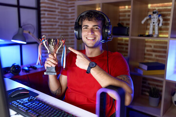 Young hispanic man playing video games holding trophy smiling happy pointing with hand and finger