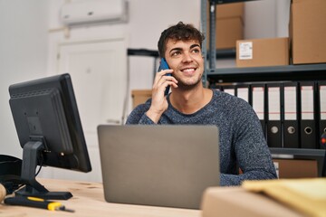 Young hispanic man ecommerce business worker using laptop talking on smartphone at office