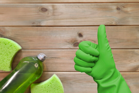 Thumb up against background of cleaning product. Hand in green glove shows like on wooden table with kitchen sponge and detergent. Selective focus. All-Natural Cleaning Solutions Concept