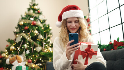 Young blonde woman using smartphone holding christmas gift at home