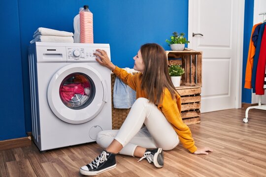 Young blonde woman sitting on floor turning washing machine on at laundry room