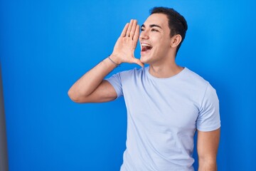 Young hispanic man standing over blue background shouting and screaming loud to side with hand on mouth. communication concept.