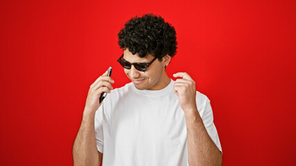 Young latin man dancing listening to music on smartphone wearing sunglasses over isolated red...
