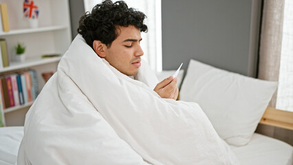 Obraz premium Young latin man covering with blanket holding thermometer at bedroom
