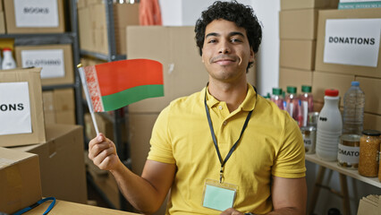 Young latin man volunteer holding belarus flag smiling at charity center - obrazy, fototapety, plakaty