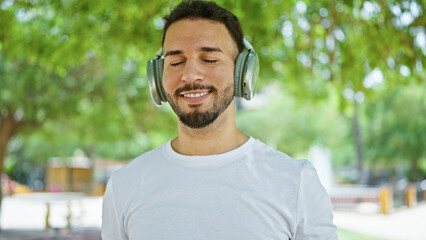 Young arab man listening to music smiling at park - obrazy, fototapety, plakaty