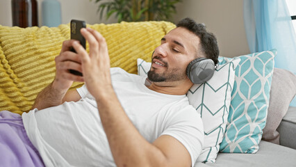 Young arab man using smartphone and headphones lying on sofa at home