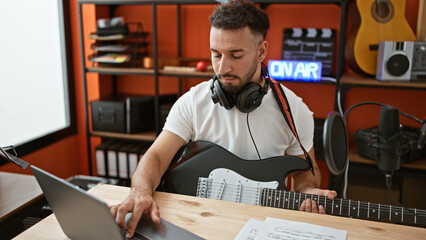 Young arab man musician playing electrical guitar composing song using laptop at music studio
