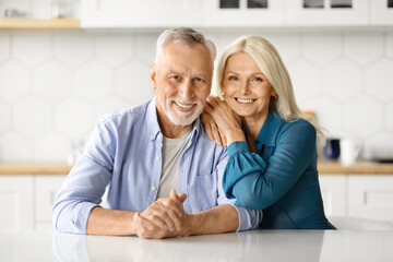 Portrait Of Happy Senior Spouses Posing At Table In Kitchen,